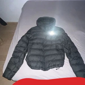 Svart dunjacka från Adidas - Säljer en svart dunjacka från Adidas med dragkedja och hög krage. Jackan är quiltad och har en sportig look, perfekt för kyligare dagar. Den har långa ärmar och en diskret logga på bröstet.