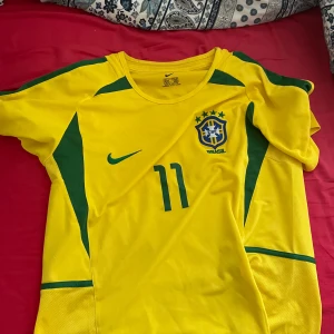 Brasilen 2002 - Säljer en gul Brasilien fotbollströja från Nike med gröna detaljer och nummer 11 på framsidan. Tröjan har korta ärmar och texten 'Ronaldinho' på baksidan. Perfekt för fotbollsfans som älskar klassiska landslagströjor!