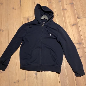 Mörkblå hoodie från Ralph Lauren - Säljer en stilren mörkblå hoodie från Ralph Lauren i storlek M med dragkedja och det klassiska logotypbroderiet på bröstet. Svin bra kvalité och bara använd ett fåtal gånger. Sjukt snygg och matchar bra med nästan alla färger.