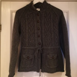 Grå stickad kofta i Lammull och angora från Boomerang  - Säljer en mysig grå stickad kofta med knappar och knytband i midjan i Lammull och angpra från Boomerang. Koftan har ett fint flätmönster och två fickor framtill. Perfekt för kyliga dagar!