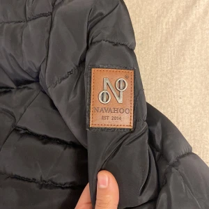 Svart pufferjacka från Navahoo - Snygg svart pufferjacka från Navahoo med quiltad design och fuskpäls i luvan. Jackan har dragkedja och knappar framtill samt praktiska fickor med dragkedjor och läderdetaljer. Perfekt för kyliga dagar!