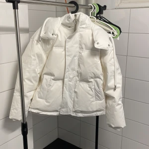 Ami Dunjacka | Vit - Helt ny och snygg vit dunjacka från Ami med dragkedja och hög krage. Nypris på jackan är ungefär 8000kr. Jackan har praktiska fickor och en broderad logotyp på ärmen. Perfekt för kyliga dagar med stil. Den har en grå fläck på brösten, använt fåtal gånger och säljes då den aldeles är för stor för mig. Jackan är i storleken M/L. Längd: ~75cm, bredd: ~60cm, längd på armen: ~65cm.