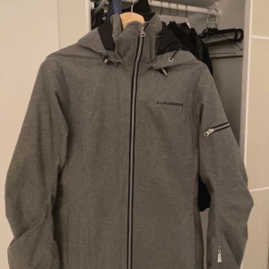 Grå skidjacka från J.Lindeberg - Snygg grå skidjacka från J.Lindeberg med dragkedja och huva. Perfekt för en kall dag i backen eller för dig som bara vill vara extra varm i vinter Jackan har en praktisk ficka med dragkedja på ärmen.