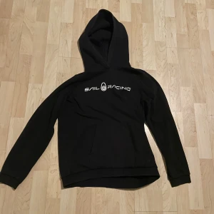 Svart hoodie från Sail Racing - Säljer en svart hoodie från Sail Racing med vit logga på bröstet. Tröjan har en klassisk design med huva och långa ärmar. Perfekt för en avslappnad stil.