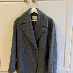 Grå ullkappa från H&M - Snygg grå kappa i ull från H&M med klassisk krage och stilren design. Perfekt för kyliga dagar med sin långa ärm och mjuka material. En tidlös favorit i garderoben.