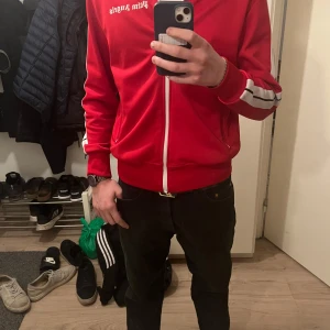 Palm angels  - Snygg röd track jacket från Palm Angels med vita ränder längs ärmarna och dragkedja framtill. Jackan har en hög krage och text på bröstet. Perfekt för en sportig look.