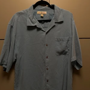 Blå silkes skjorta från Tommy Bahama - Säljer en stilren blå skjorta från Tommy Bahama i 100% silke. Skjortan har korta ärmar och en klassisk krage med knappar framtill. Perfekt för en avslappnad och elegant look.Klippt snyggt så den är bra längd 