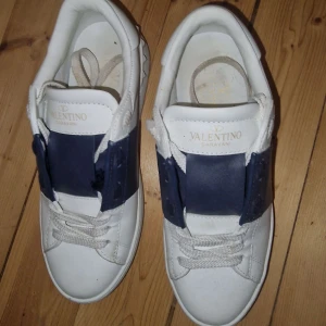 Valentino Garavani sneakers i vitt och blått - Snygga Valentino Garavani sneakers i vitt med en bred blå rem över snörningen. Skorna har en stilren design med klassiska snören och en bekväm sula. Perfekta för en trendig look. Storlek 38