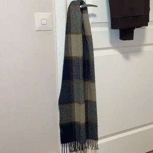 Rutig halsduk från Tommy Hilfiger - Snygg rutig halsduk från Tommy Hilfiger i blått och beige. Perfekt för att hålla dig varm och stilren under kyliga dagar. Den har fransar i ändarna. 