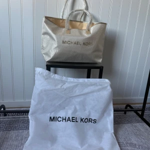 Glittrig väska från Michael Kors - Snygg och glittrig väska från Michael Kors i en ljus färg. Väskan har två handtag och är perfekt för att ge en touch av glamour till din outfit. Den har en stilren design med Michael Kors-loggan på framsidan.