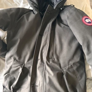 Grå chateau parkajacka från Canada Goose - Säljer en stilren grå parkajacka från Canada Goose med deras ikoniska märke på ärmen. Jackan har en dragkedja och knappar framtill samt praktiska fickor. Perfekt för kalla dagar. Slitningar vid båda handlederna och ett litet hål som på bilden annars bra, (Ingen päls)
