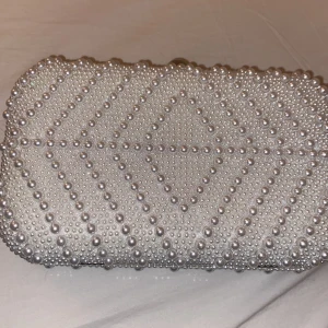 Vit pärlbeströdd kuvertväska - Elegant vit kuvertväska dekorerad med pärlor i ett geometriskt mönster. Väskan har en metallram med en rund knäppning och en svart insida. Perfekt för speciella tillfällen.