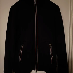 Svart jacka med dragkedja från Moncler - Snygg svart jacka från Moncler med dragkedja och två fickor framtill. Jackan har en hög krage och Moncler-logga på ärmen. Perfekt för en stilren look. Storlek S/M passar dig slm är 178-185 har ett hål på armen som ör lagat och syns knappt