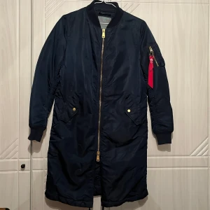Mörkblå lång bomberjacka från Alpha Industries S - Snygg mörkblå bomberjacka från Alpha Industries med gulddragkedja och ribbade muddar. Jackan har en lång passform och är quiltad på insidan för extra värme. Perfekt för höst och vinter.