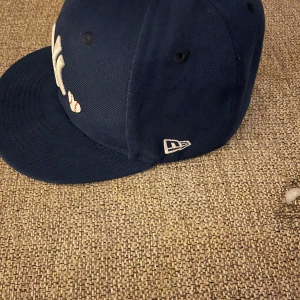 Mörkblå keps från New Era - Snygg mörkblå keps från New Era med broderad logga framtill och World Series-märke på sidan. Perfekt för sportiga tillfällen eller som en stilren accessoar.