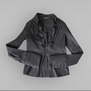 Svart blus med volangdetaljer från kiwi - Elegant svart blus från kiwi med volangdetaljer längs kragen och knappar framtill. Långärmad design med knappar vid ärmsluten för en stilren look. Perfekt för en chic och sofistikerad stil.