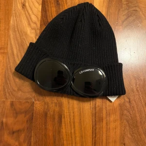 Svart mössa med glasögon från C.P. Company - Snygg svart ribbad mössa från C.P. Company med inbyggda glasögon på uppviket. Perfekt för att ge en unik touch till din outfit.