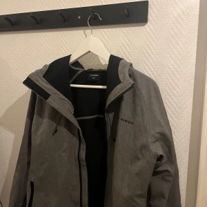 Grå vindjacka från Everest - Snygg grå vindjacka från Everest med dragkedja och justerbar huva. Perfekt för blåsiga dagar och utomhusaktiviteter. Jackan har en stilren design med diskret logga på bröstet. Funkar bra vardagligt och till skidåkning ✅ M