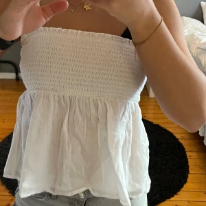 Vit axelbandslös topp - Säljer en söt vit axelbandslös topp med smockdetaljer upptill och en luftig passform nedtill. Kan användas både som kjol och topp