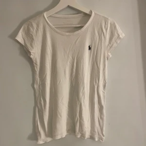 Vit t-shirt från Ralph Lauren - Säljer en klassisk vit t-shirt från Ralph Lauren med korta ärmar och rund halsringning. Den har en liten broderad logotyp på bröstet. Perfekt för en stilren och enkel look.