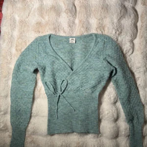 Grön omlottstickad tröja från Urban Outfitters - Säljer en mysig grön omlottstickad tröja från Urban Outfitters. Tröjan har en v-ringad design med knytband i midjan och långa ärmar. Perfekt för kyliga dagar!