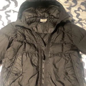 Svart Stone Island jacka  - Säljer en svart dunjacka från Stone Island med huva och dragkedja. Jackan har en broderad logotyp på ärmen och praktiska fickor med knappar framtill. Perfekt för kyliga dagar. Orginal pris 10 000