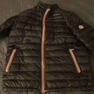 Svart dunjacka från Moncler - Snygg svart dunjacka från Moncler med quiltad design och färgglada dragkedjor. Perfekt för kyligare väder med sin lätta och varma konstruktion. Jackan har långa ärmar och en klassisk passform.