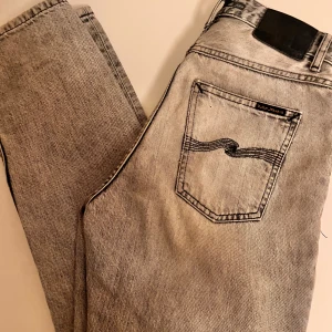 Grå jeans från Nudie Jeans - Passar 29/32Snygga grå jeans från Nudie Jeans med en klassisk femficksdesign och knappgylf. De har en stilren look med en diskret tvättad effekt. Perfekta för en avslappnad stil.