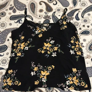Svart blommig linnetopp - Säljer en superfin svart linnetopp med blommigt mönster i gult och vitt. Toppen har smala axelband och en v-ringad hals. Perfekt för sommardagar eller en avslappnad festkväll. 🌼 Den är lite genomskinlig så topp kan behövas.