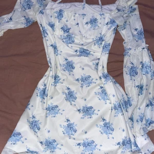 Vit och blå blommig klänning - Säljer en superfin vit klänning med blå blommor. Den har en söt offshoulder-design med långa ärmar och en smickrande passform. Perfekt för sommarens fester eller en mysig vårdag. Klänningen har en romantisk stil med volangdetaljer vid ärmsluten.