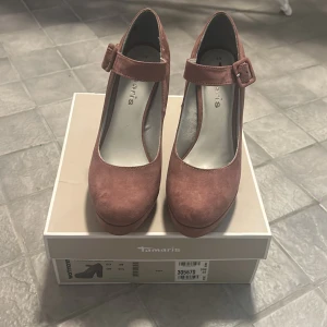 Mauve klackskor från Tamaris - Säljer ett par snygga mauvefärgade klackskor från Tamaris. De har en elegant rem med spänne över foten och en stabil klack som ger bra stöd. Perfekta för både fest och vardag!