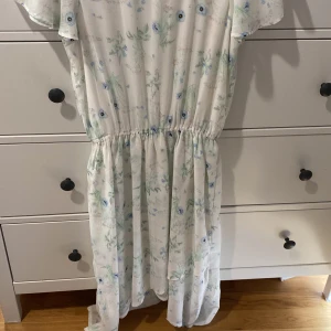 Vit blommig klänning - Säljer en superfin vit klänning med blommigt mönster i ljusblått och grönt. Oanvänd, fortfarande ny. Klänningen har korta ärmar och en bekväm passform med resår i midjan. Perfekt för sommarens alla fester eller en mysig dag i parken! Köpt för 300kr