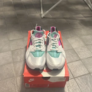 Nike Air Huarache i vit, turkos och rosa - Säljer ett par snygga Nike Air Huarache sneakers i en fräsch färgkombination av vit, turkos och rosa. Skorna är i nyskick och kommer i originalkartong. Obs, denna model är stor i storleken och passar dig som vanligtvis har storlek 38,5 eller 39. 