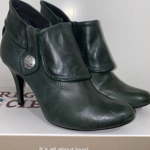 Gröna skinnstövletter med klack - Säljer ett par eleganta gröna stövletter i skinn med en snygg klack. De har en dekorativ knappdetalj på sidan och en spetsig tå som ger en stilren look. Perfekta för höst och vinter, både till vardags och fest.