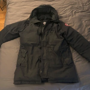 Ganada goose jacka - Grym vinterjacka, håller värmen bra och praktisk med många fickor! Använd ett två säsonger varsamt! Skicket är okej.  En knapp har tappat fäste, borde vara lätt att sy fast för den som kan.  