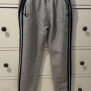 Grå mjukisbyxor från Adidas - Säljer ett par grå mjukisbyxor från Adidas. De har en bekväm passform med elastisk midja och två sidofickor. Passar för både tjejer och killar. Använd en gång. Går inte till användning längre då storleken inte passade mig
