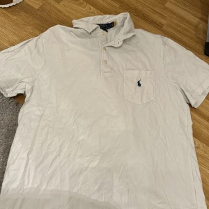 Vit kortärmad T-shirt pikétröja från Ralph Lauren - Säljer en klassisk vit T-shirt pikétröja från Ralph Lauren i bomull. Den har en broderad logga på bröstfickan och knappar vid kragen. Perfekt för en avslappnad men stilren look. Passar bra till både vardag och fest!