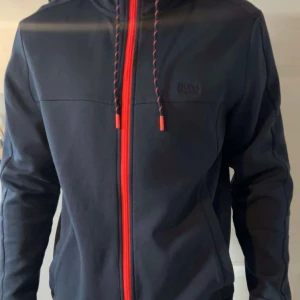 Marin blå hoodie från hugo boss, storlek M - Säljer en marinblå hoodie med rosa detaljer från hugo boss i storlek m. Använd endast ett par gånger, nästan i nyskick. 