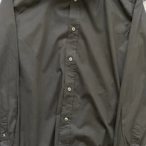 Svart skjorta från Dolce & Gabbana - Säljer en elegant svart skjorta från Dolce & Gabbana, tillverkad i Italien. Skjortan är i bomull med en liten del elastan för extra komfort. Den har långa ärmar och knappar framtill. Perfekt för både formella och avslappnade tillfällen. Perfekt för fest osv.