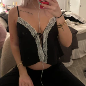 Svart linne med spetsdetaljer - Säljer ett svart linne med vita spetsdetaljer och knytning framtill. Toppen är ärmlös och har smala axelband, perfekt för en somrig look eller en utekväll. Den är både söt och lite edgy med sin kontrast mellan svart och vit spets.