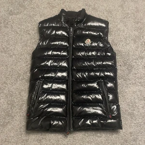 Svart dunväst från Moncler - Säljer en snygg svart dunväst från Moncler i mycket bra skick. Västen har en glansig finish och är perfekt för höst och vinter. Den har en dragkedja framtill och två praktiska fickor med dragkedjor. Moncler-loggan sitter på bröstet. Perfekt för att hålla sig varm och stilren! Har änvänt Max 2 gånger. Storlek 1 asså S