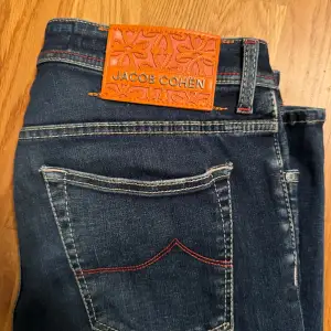 Säljer ett par snygga mörkblå jeans från Jacob Cohen i storlek 33. De har en klassisk femficksdesign med röda sömmar och en cool orange läderpatch med logga baktill. Jeansen har knappar istället för dragkedja och en broderad detalj på myntfickan. Perfekta för både vardag och fest! Bra skick förutom en defekt utpekad med röd pil.