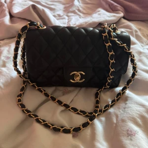 Svart quiltad axelväska - Säljer en svart axelväska med guldfärgade kedjedetaljer. Väskan har en klassisk design med en snygg logotyp på framsidan och en praktisk innerficka. Perfekt för både vardag och fest! 700kr kan gå ner till 500kr.