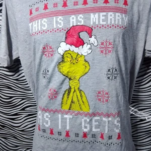 Grå jultröja med Grinch-motiv - Säljer en grå t-shirt med ett roligt Grinch-motiv och texten 'This is as merry as it gets'. Perfekt för julen med sitt festliga tryck i rött och grönt. T-shirten är kortärmad och i bomull, vilket gör den bekväm att bära hela dagen. Passar perfekt för en avslappnad julfest eller som en rolig present!