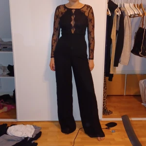 Svart jumpsuit med spetsdetaljer - Snygg svart jumpsuit med långa spetsärmar och transparent rygg. Perfekt för festliga tillfällen! Den har en tight passform upptill och vida byxben. Dragkedja i ryggen för enkel påklädning.