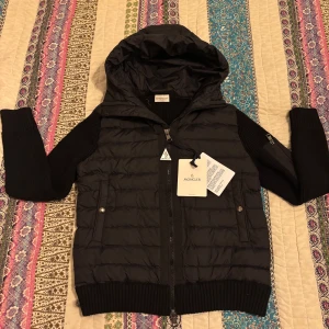 Moncler Padded Wool Cardigan - Säljer en snygg svart jacka från Moncler i ny skick. Jackan har en quiltad design med dragkedja och huva. Den är perfekt för höst och vår, passar perfekt för en stilren look!