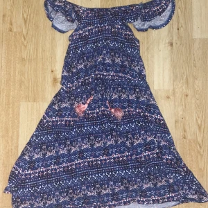 Blå offshoulder klänning med mönster - Säljer en superfin blå offshoulder klänning med ett färgglatt mönster. Klänningen har volangdetaljer vid axlarna och söta tofsar framtill. Perfekt för sommarens fester eller en dag på stan. Materialet känns lätt och luftigt, vilket gör den idealisk för varma dagar. Passar perfekt för en bohemisk stil!
