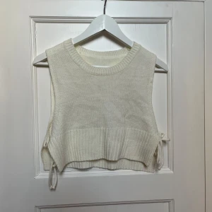 Vit stickad väst - Säljer en superfin vit stickad väst i croppad stil. Den har ribbade kanter och snygga knytband på sidorna för en extra touch. Perfekt för lager-på-lager under hösten eller våren. Passar till både jeans och kjol!