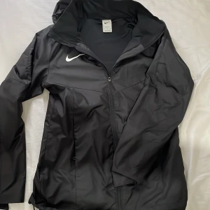 Svart vindjacka från Nike - Säljer en svart vindjacka från Nike i mycket bra skick. Jackan har en dragkedja framtill och en praktisk huva. Perfekt för höst och vår, tillverkad i polyester för att stå emot vind och lättare regn. Den har en stilren design med Nike-loggan på bröstet. Passar både till träning och vardags.