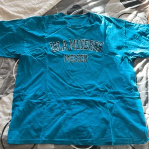 Turkos t-shirt med Isla Mujeres-tryck - Säljer en turkos t-shirt med texten 'Isla Mujeres Mexico' tryckt på framsidan. Perfekt för en avslappnad stil eller som souvenir. T-shirten är i bra skick och har en normal passform. Perfekt för sommardagar eller som en påminnelse om soliga stränder.